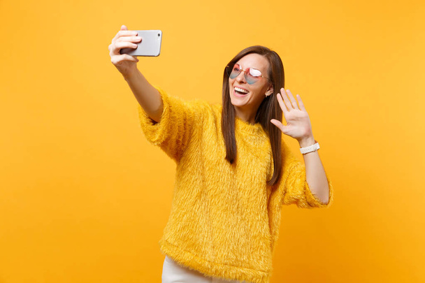 Smiling woman in heart glasses doing taking selfie shot on mobile phone making video call waving hand for greeting isolated on bright yellow background. People sincere emotions, lifestyle. Copy space - Zdjęcie, obraz