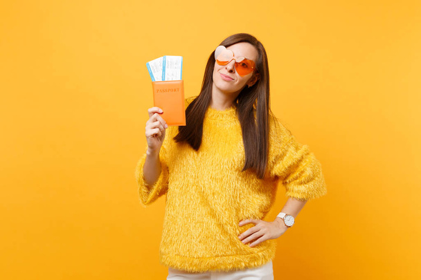 Привлекательная молодая женщина в меховом свитере, оранжевые очки сердца с паспортом и посадочными талонами изолированы на ярко-желтом фоне. Люди искренние эмоции, образ жизни. Рекламная зона
 - Фото, изображение