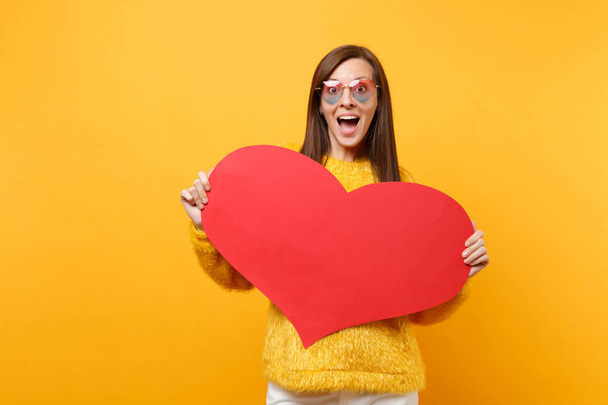 Portret van opgewonden verbaasd jonge vrouw in bont trui hart brillen houden lege lege rood hart geïsoleerd op heldere gele achtergrond. Mensen oprechte emoties, lifestyle concept. Gebied van de reclame - Foto, afbeelding