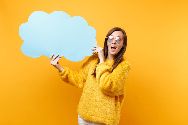 Excited shocked young woman in heart eyeglasses holding empty blank blue Say cloud, speech bubble isolated on bright yellow background. People sincere emotions, lifestyle concept. Advertising area - Photo, image