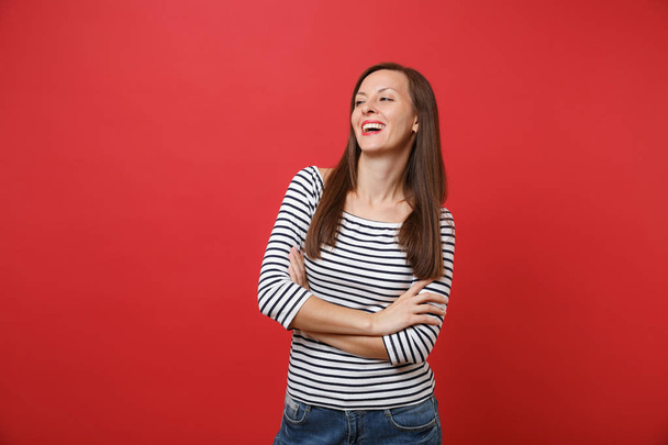 Portrait of laughing young woman in striped clothes standing, looking aside holding hands folded isolated on bright red wall background. People sincere emotions, lifestyle concept. Mock up copy space - Photo, Image