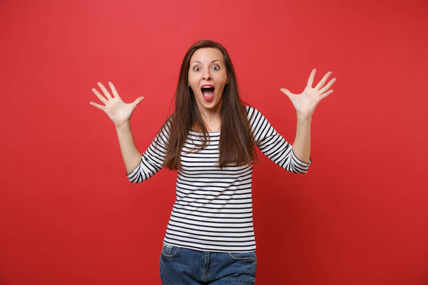 Portrait of shocked young woman keeping mouth wide open, looking surprised and spreading hands isolated on bright red wall background. People sincere emotions, lifestyle concept. Mock up copy space - Φωτογραφία, εικόνα