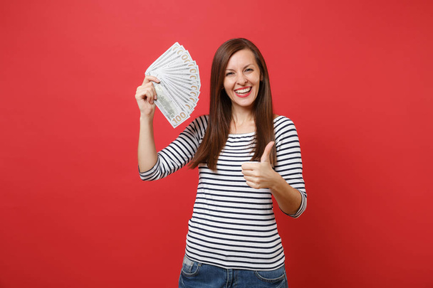 Portrait of joyful young woman showing thumb up and holding bundle lots of dollars, cash money isolated on bright red wall background. People sincere emotions, lifestyle concept. Mock up copy space - Фото, зображення