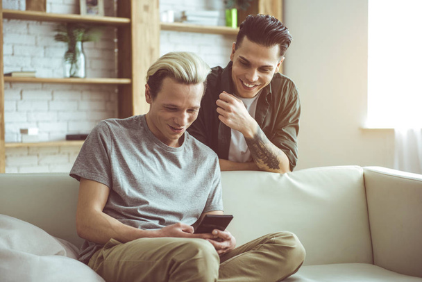 Surfing the internet. Toned portrait of handsome blond guy sitting on couch and holding smartphone while his boyfriend standing behind - Photo, image