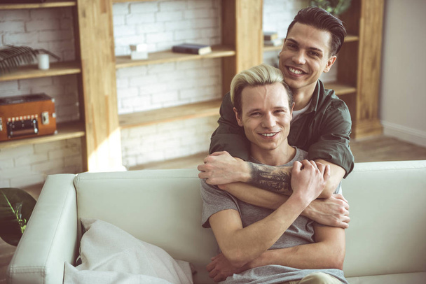 Young blond man sitting on couch while his boyfriend hugging him from behind - Фото, зображення
