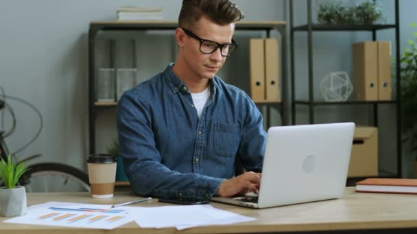 Attractive young business man in the glasses working in the modern office using laptop, looking to the camera and smiling. - Кадры, видео