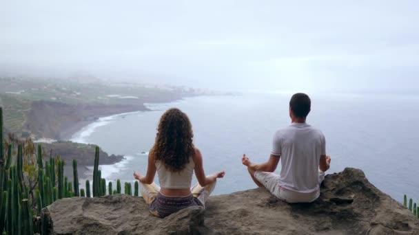 Een man en een vrouw zit op de top van een berg kijken naar de oceaan zittend op een steen mediteren in een Lotus-positie. Het uitzicht vanaf de achterkant. Canarische eilanden - Video