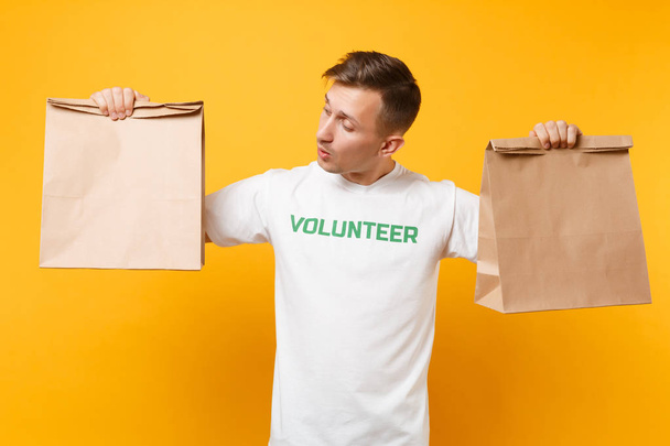 Portrait of man in white t-shirt written inscription green title volunteer hold blank craft paper bag for takeaway isolated on yellow background. Voluntary free assistance help charity grace concept - Foto, imagen
