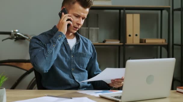 attraktiver junger Geschäftsmann mit Brille, der mit Partner auf dem Smartphone telefoniert, während er Dokumente checkt. - Filmmaterial, Video