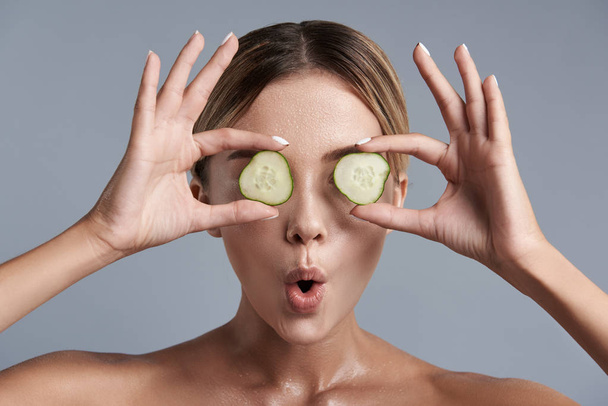 Expressive woman making funny faces while holding pieces of cucumbers - Foto, afbeelding