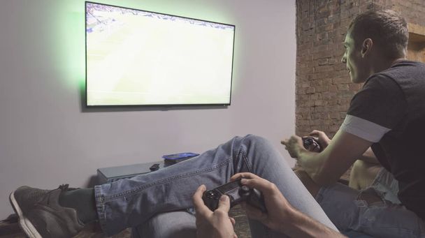 Two men playing video game Pro Evolution Soccer. Hands holding console controller. Football or soccer game on the television. Widescreen tv hang on the wall - Fotó, kép