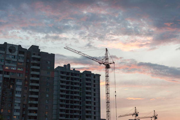 Evening at the construction site. Lifting crane builds a tall multi-storey house against a red cloudy sky in the rays of the setting su - Valokuva, kuva