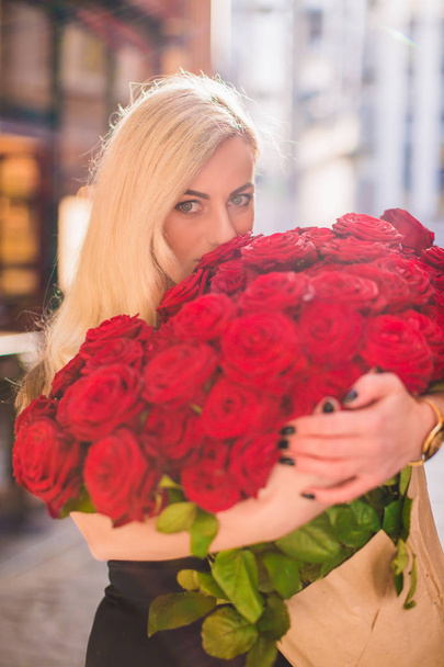 Attractive blonde woman keeps big bouquet of red roses in hands and sniffs it, on the street - Foto, Imagen