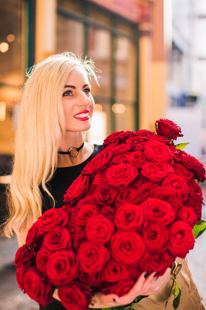 Attractive smiling lady stands and keeps big bouquet of red roses in hands, on the street - Фото, зображення
