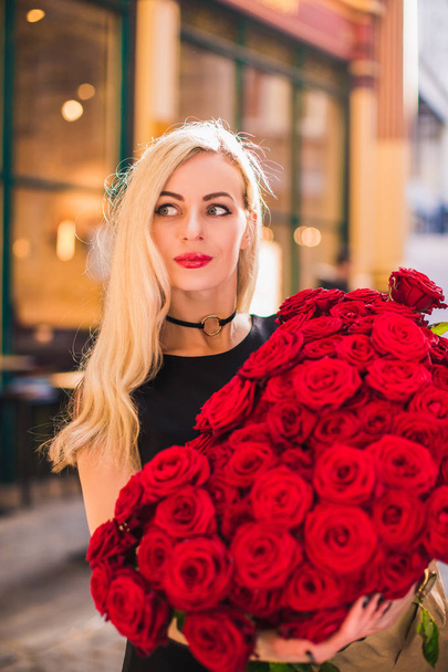 Divine lady stands and keeps big bouquet of red roses in hands, looks aside, on the street - Fotografie, Obrázek