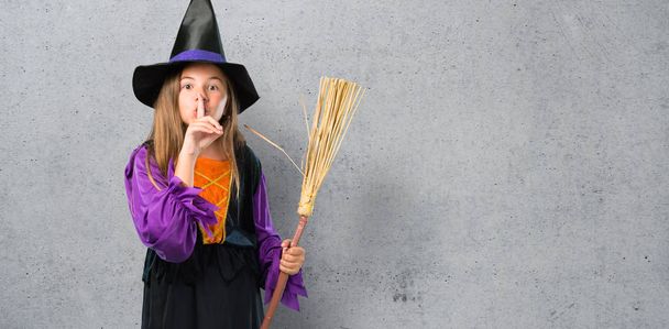 Маленька дівчинка, одягнена як відьма для святкових днів на Хеллоуїн, показує знак закриття рота і жесту мовчання на текстурованому фоні
 - Фото, зображення