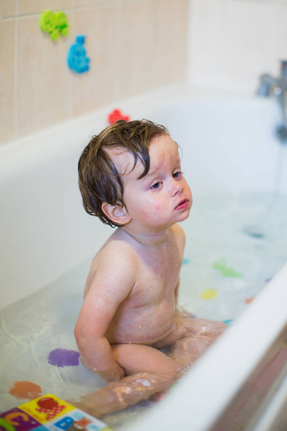 Weak little boy sits sick on rashes in bath with water and toys, indoors - Foto, afbeelding