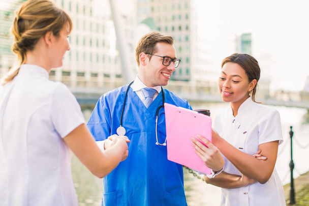 Young handsome doctor shakes hand of nurse and shows folder to another nurse outdoor - Foto, immagini