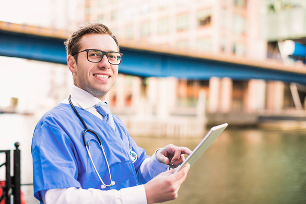 Close-up handsome smiling doctor keeps the tablet and stands against the background of the river and buildings, outdoors - Photo, image