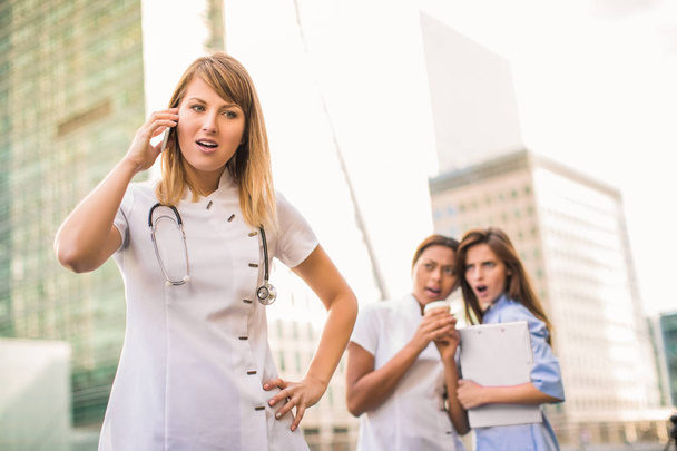 Красивий лікар стоїть і говорить по телефону на тлі двох молодих здивованих медсестер, які дивляться на лікаря, на відкритому повітрі
 - Фото, зображення