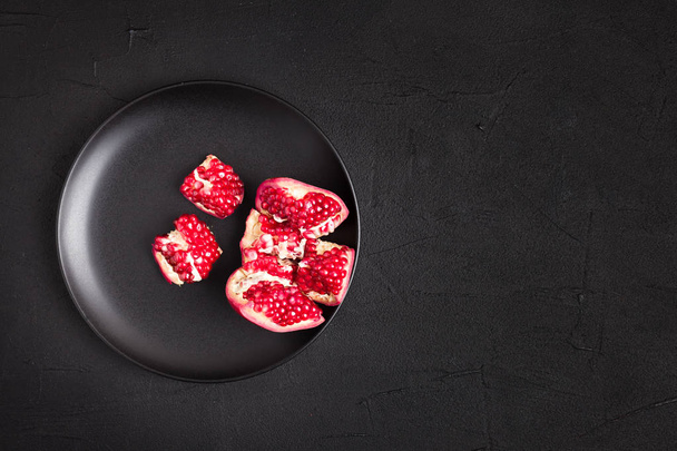 reife rot geöffnete Granatapfel-Nahaufnahme auf schwarzem Teller auf dunklem Hintergrund mit Kopierraum - Draufsicht auf saisonal roh geschnittene saftige Früchte mit Kernen in dunklem Stimmungsstil mit leerem Platz für Text. - Foto, Bild