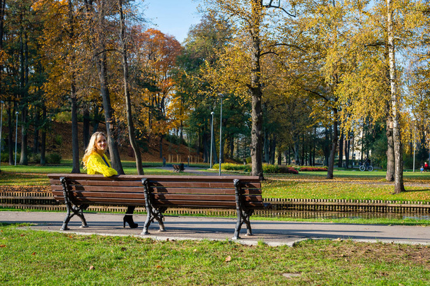 A young, beautiful, blonde woman sitting in a park on a bench. - Фото, зображення