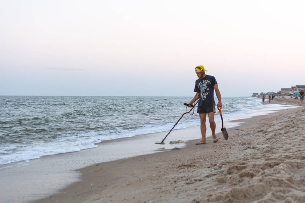 Caçador de tesouros com um detector de metais caminha ao longo da praia do Mar Negro após o pôr do sol à procura de tesouros
 - Foto, Imagem