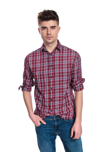портрет расслабленного молодого человека в рубашке с пледами, стоя на белом фоне с руками в карманах
 - Фото, изображение