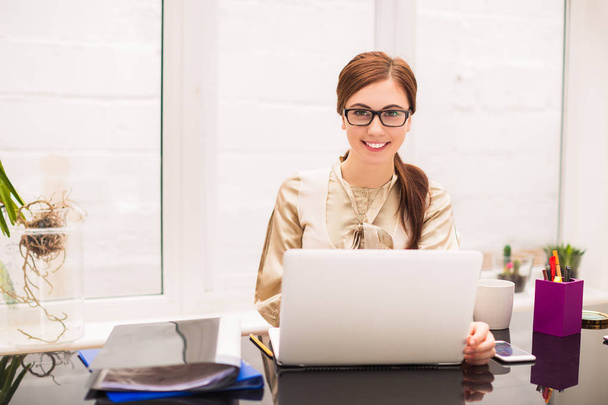 Young cheerful business woman sits at the table with laptop and folders at the office - Foto, Imagen