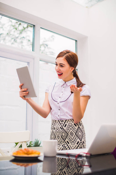 Young beautiful business woman keeps the tablet in hand, looks at it and smiles at the office - Fotoğraf, Görsel