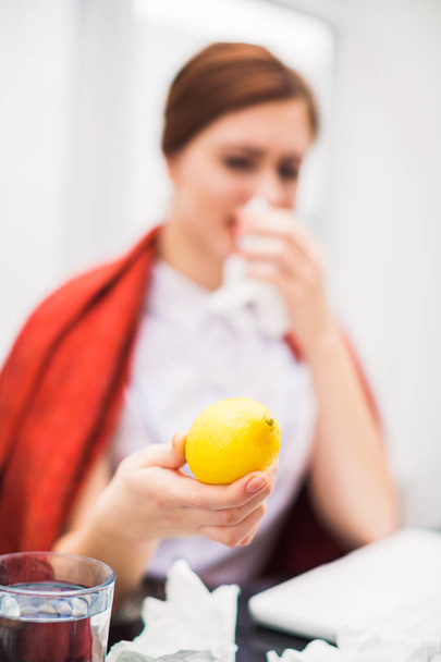 Young sick office manager sits at the table at the office  with napkins in hand, wipes nose and keeps a lemon in hand - Foto, Imagen