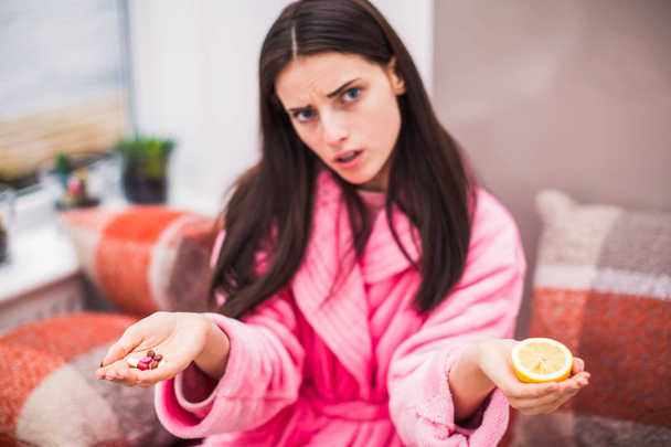 Portrait of young sick woman who sits on bed at home and keeps pills and lemon in hands - 写真・画像