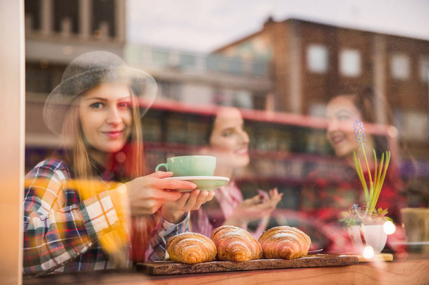Foreground woman with cup of coffee in her hands and croissants on the table, background two women tell cute, in a cafe - 写真・画像