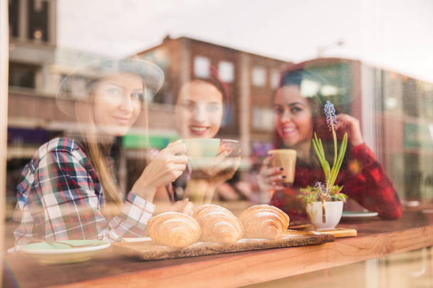 Three beautiful women sit at the table with croissants and cups of coffee in their hands, in a cafe - Photo, image
