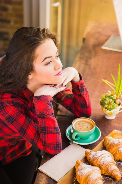 Beautiful woman sits at the table with the croissants and cup of coffee and looks in window in cafe - 写真・画像