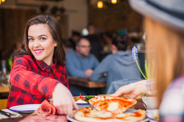 zwei Studentinnen sitzen am Tisch mit Pizza und anderen Lebensmitteln, sie nehmen Pizza in Scheiben, in einer Pause, im Restaurant - Foto, Bild