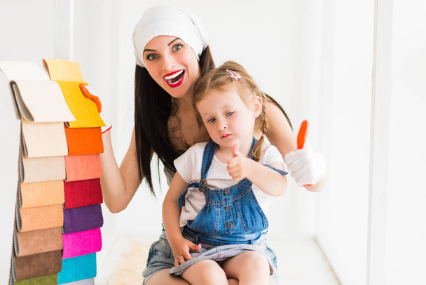 Portrait of young happy woman who keeps her daughter on lap and keeps color palette in hand indoors - Foto, Imagen