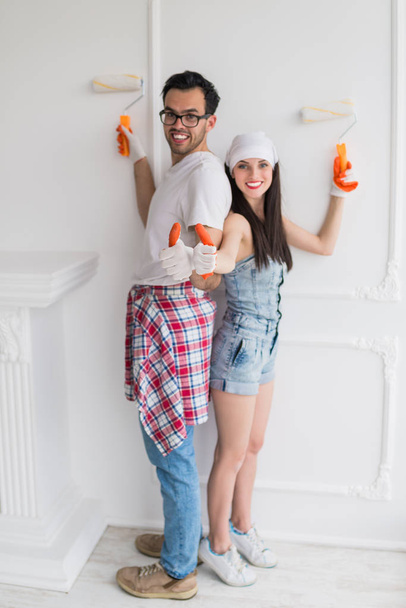 The young joyful couple does repair at home, they paint the wall with help of platens and keep thumbs up - Foto, Imagem