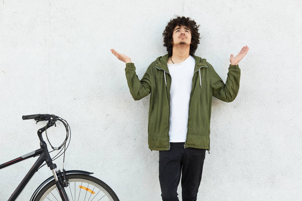 Oh God, help me please! Plesding male teenager spreads palms and raises head, prays for something while stands against white background near bike. People, body language and lifestyle concept - Photo, image