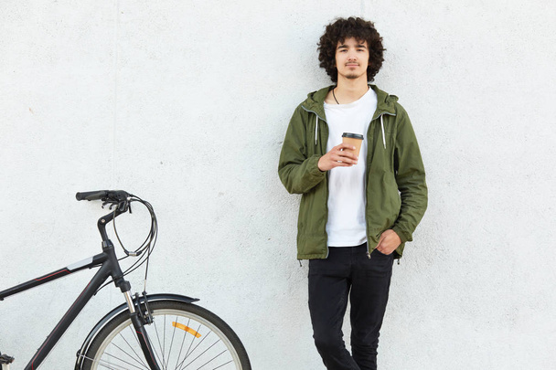 Indoor shot of handsome guy with curly hair, dressed in fashionable jacket, black trousers, drinks hot beverage, stands near bicycle against white concrete wall. People, cycling and lifestyle concept - Photo, image