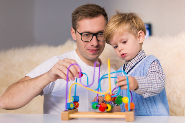 The little cute serious son playing by developing toy near his young happy father indoors - Фото, зображення
