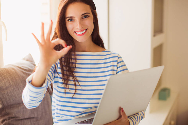 Portrait of young cheerful bewitching woman who sitting on the sofa in the room and keeping laptop and showing sign "cool" her hands - Фото, зображення