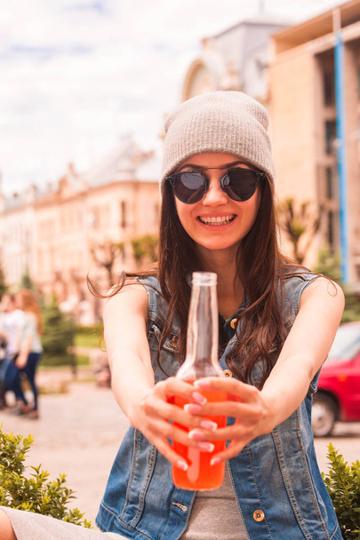 Portrait of young stylish woman who sitting outdoor and smiling and keeping bottle of juice in hands - Фото, изображение