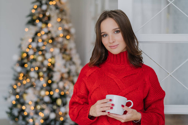 Красива жінка з приємним зовнішнім виглядом і макіяжем, носить теплий в'язаний зимовий светр, тримає чашку кави, стоїть біля прикрашеної новорічної ялинки, планує відсвяткувати свято. Концепція відпочинку
 - Фото, зображення