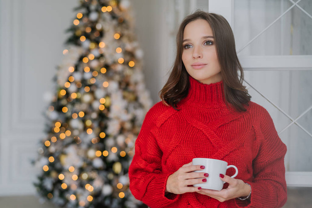 Plan intérieur d'une jeune femme réfléchie en vêtements d'hiver chauds, apprécie le café chaud ou le cappuccino, regarde pensivement de côté, se tient près de l'arbre de Noël décoré, a une manucure rouge. Concept de confort
 - Photo, image