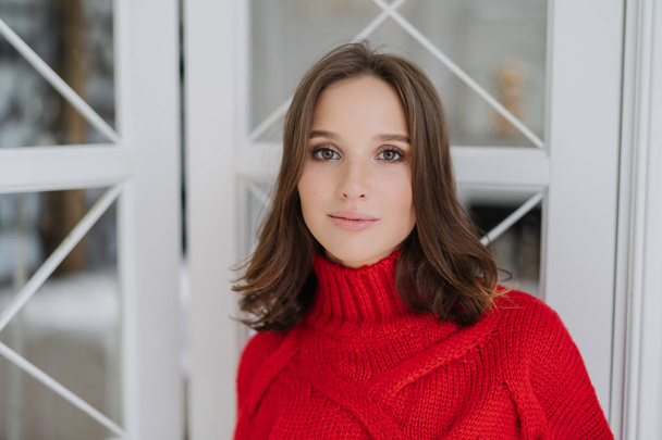 Horizontal shot of calm beautiful young European woman wears warm red sweater, has make up and healthy soft skin, poses indoor, spends spare time at home. People, beauty and lifestyle concept - Photo, Image