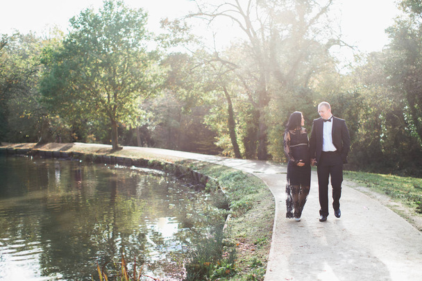 A young pregnant woman walks near pond in the park with her happy husband and they keep hands each other - Foto, Imagem