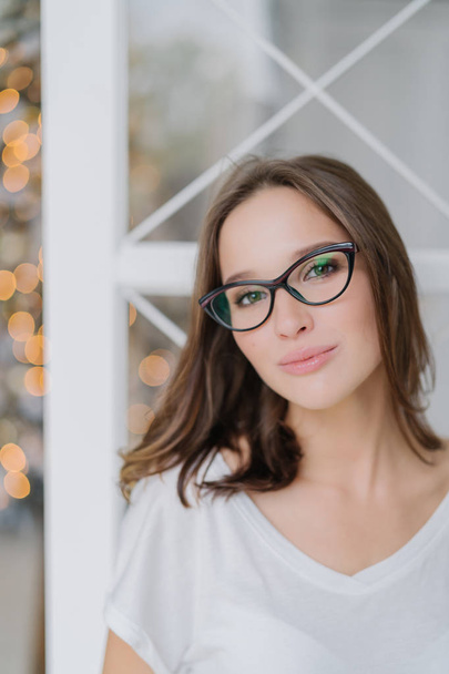 Vertical shot of charming woman in optical glasses, dressed casually, poses indoor against Christams tree, has healthy soft skin, enjoys spare time in domestic atmosphere. People and leisure - Foto, imagen