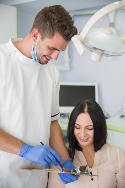 Portrait of friendly male dentist holding in his hand a clipboard and consulting with his beautiful patient at dental office. - Фото, изображение