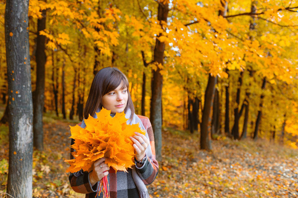 Portrait of young girl outdoor. Bouquet with yellow leafs in her hands. Brunette woman in autumn park with fashionable plaid coat and scarf. - Photo, Image